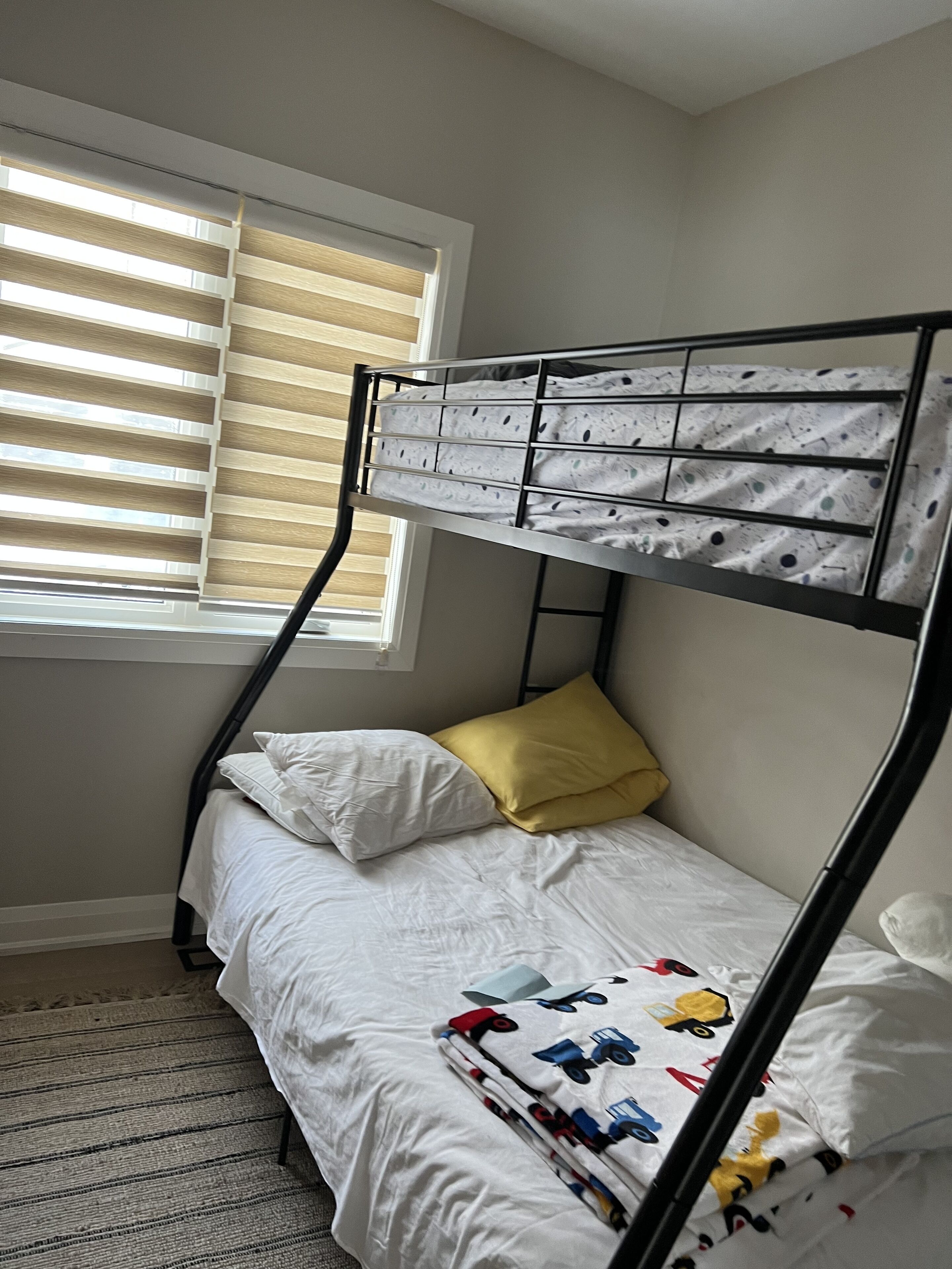 Ein Stockbett mit weißer Bettwäsche und einem gelben Kopfkissen, ein Fenster mit Jalousien und einer gemusterten Decke.