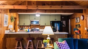 A cozy cabin interior with a wooden ceiling, a kitchen with a microwave and refrigerator, a bar area with stools, and a blue sofa with a checkered pillow. - The Bunkhouse 1960's Camp Vibe + Timeless Wet Bar (Springville)