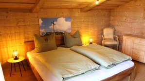 Un dormitorio de cabaña de madera con dos camas, una silla, una mesita con una lámpara y un cuadro en la pared.