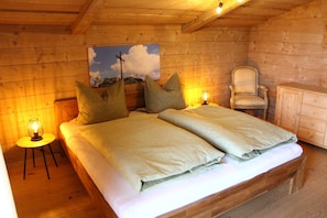 A wooden bedroom with a bed, two bedside tables, a chair, and a painting on the wall.