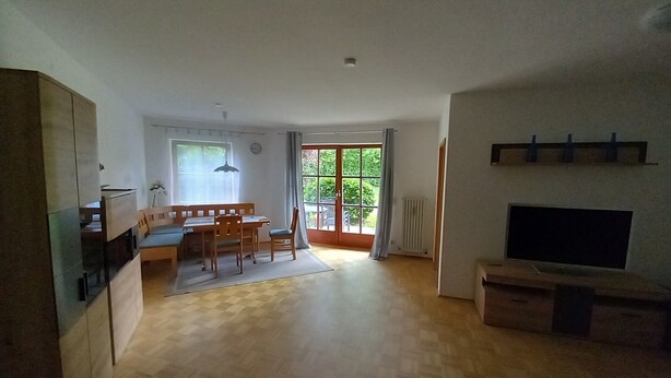 Een ruime woonkamer met een houten eettafel en stoelen, een bank, een televisie en een groot raam met uitzicht op groen.