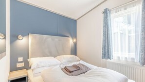 A neatly made bed with white linens, a bedside table with a lamp, and a window with curtains. - Spacious and luxurious 12ft-wide caravan with one double and one twin. (Axminster)