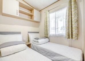 A bedroom with two beds, a window with curtains, a bedside table, and a built-in shelf above the beds. - Spacious and luxurious 12ft-wide caravan with one double and one twin. (Axminster)