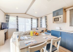 A compact, well-lit room with a dining table, chairs, a sofa, a TV, a kitchen area with cabinets and a microwave, and large windows with curtains.