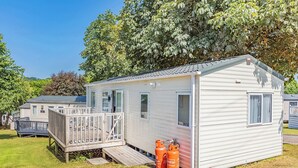 A single-story mobile home with a deck, a small porch, and propane tanks. - Spacious and luxurious 12ft-wide caravan with one double and one twin. (Axminster)