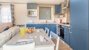 A compact kitchen with a microwave, stove, and sink. There's a dining table with chairs and a bowl of fruit. - Spacious and luxurious 12ft-wide caravan with one double and one twin. (Axminster)