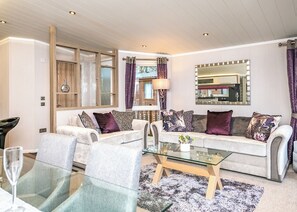 A modern living room with a glass coffee table, two sofas with patterned cushions, a large mirror, and a window with curtains.