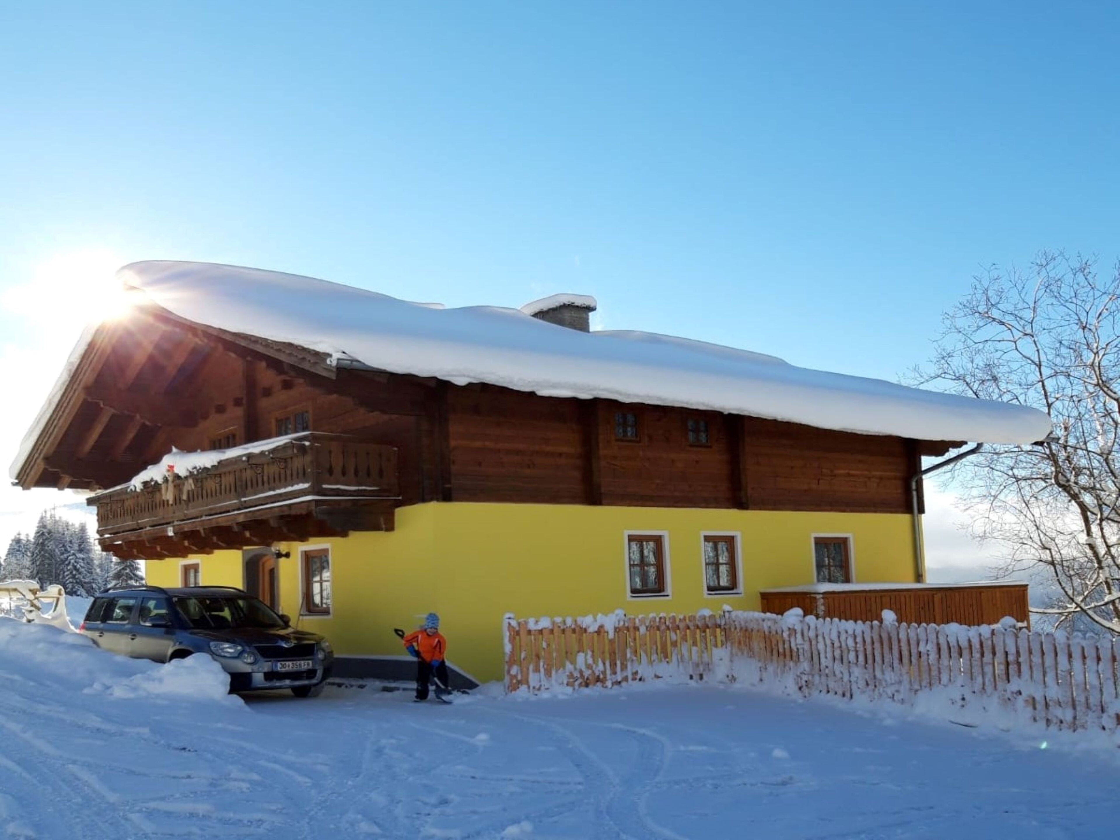 A wooden chalet with a sloped roof covered in snow, a yellow facade, and a balcony.