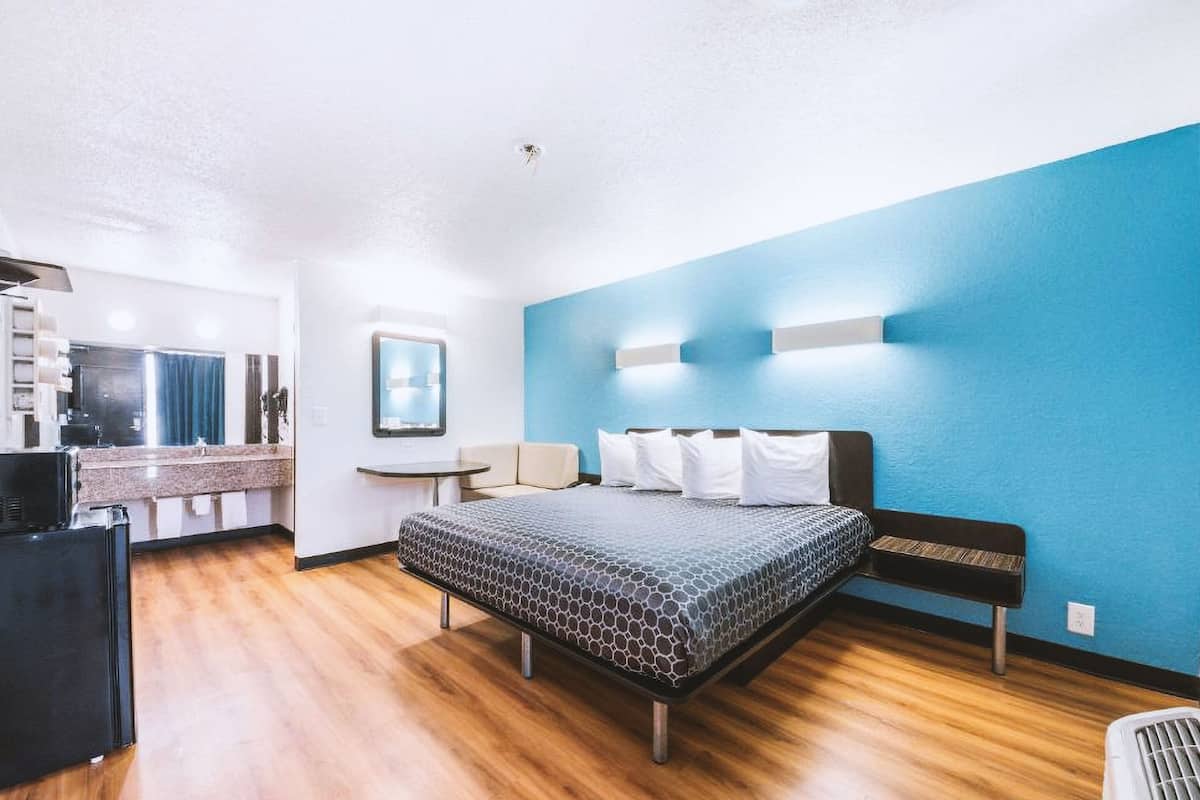 A modern hotel room with a bed, a small desk, a chair, and a kitchenette.