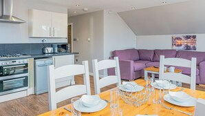 A modern kitchen with white cabinets, a dining area with a wooden table set for two, and a purple sofa in the living area.