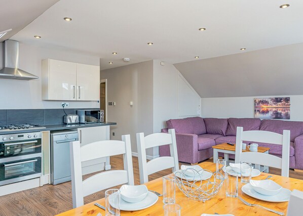 Una cocina moderna con armarios blancos, un comedor con mesa de madera para dos personas y un sofá púrpura en la sala de estar.