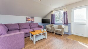 A living room with a purple sofa, a wooden coffee table, two armchairs, a TV, and a window with curtains.