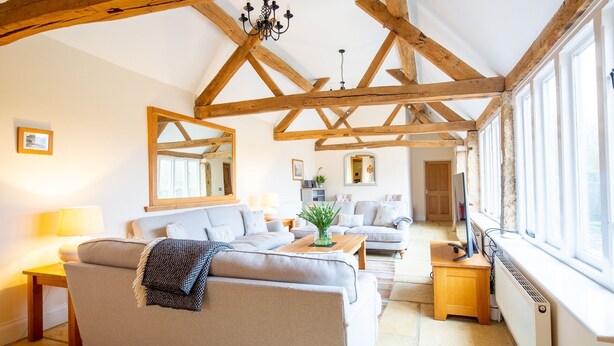 A spacious living room with exposed wooden beams, a sofa, a coffee table, and a large window providing natural light. - Detached Cotswold converted Barn near Chipping Campden (gloucestershire)