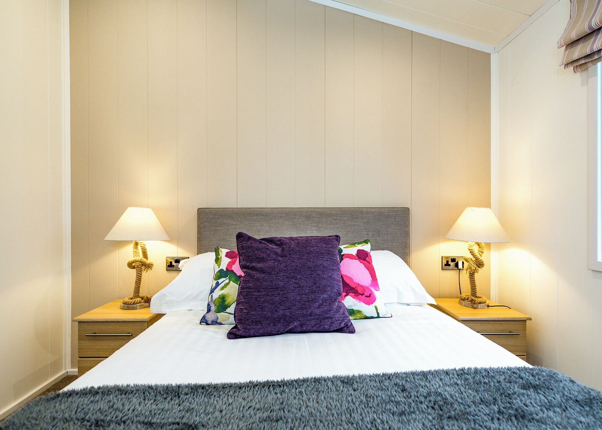 Uma cama bem arrumada com um travesseiro roxo, duas luminárias de cabeceira e uma mesinha de cabeceira de madeira.
