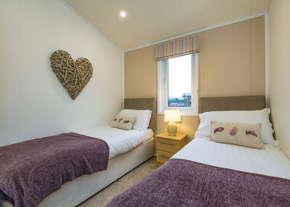 A bedroom with two beds, a nightstand with a lamp, a window with curtains, and a decorative heart on the wall.