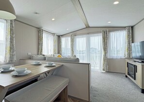 A modern hotel room with a dining area, a sofa, a TV, and large windows with curtains.