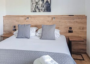 A hotel room with a wooden headboard, a large bed with white bedding, two gray pillows, and a gray throw blanket.