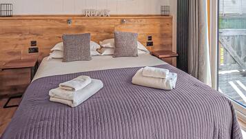 A hotel room with a wooden headboard, a bed with a purple quilt, two pillows, and two towels on the bed.