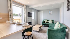 Un salon moderne avec un canapé vert, une table à manger en bois avec des chaises, un téléviseur fixé au mur et une vue sur l’extérieur.