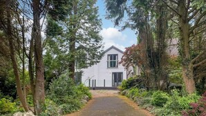 A white house with a central door, surrounded by dense greenery and trees. - 3 bedroom accommodation in Milnthorpe (Milnthorpe)