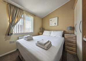 Un dormitorio con cama, mesita de noche, cómoda, ventana con cortinas y lámpara de pared.