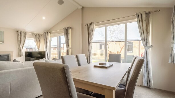A modern living room with a sofa, a dining table, and chairs. There is a TV on a stand, and large windows with curtains.