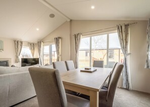 A modern living room with a sofa, a dining table, and chairs. There is a TV on a stand, and large windows with curtains.