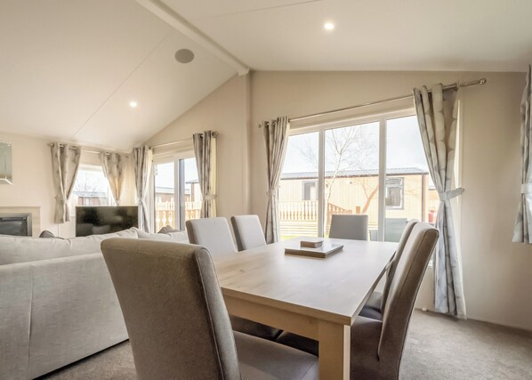 A modern living room with a sofa, a dining table, and chairs. There is a TV on a stand, and large windows with curtains.