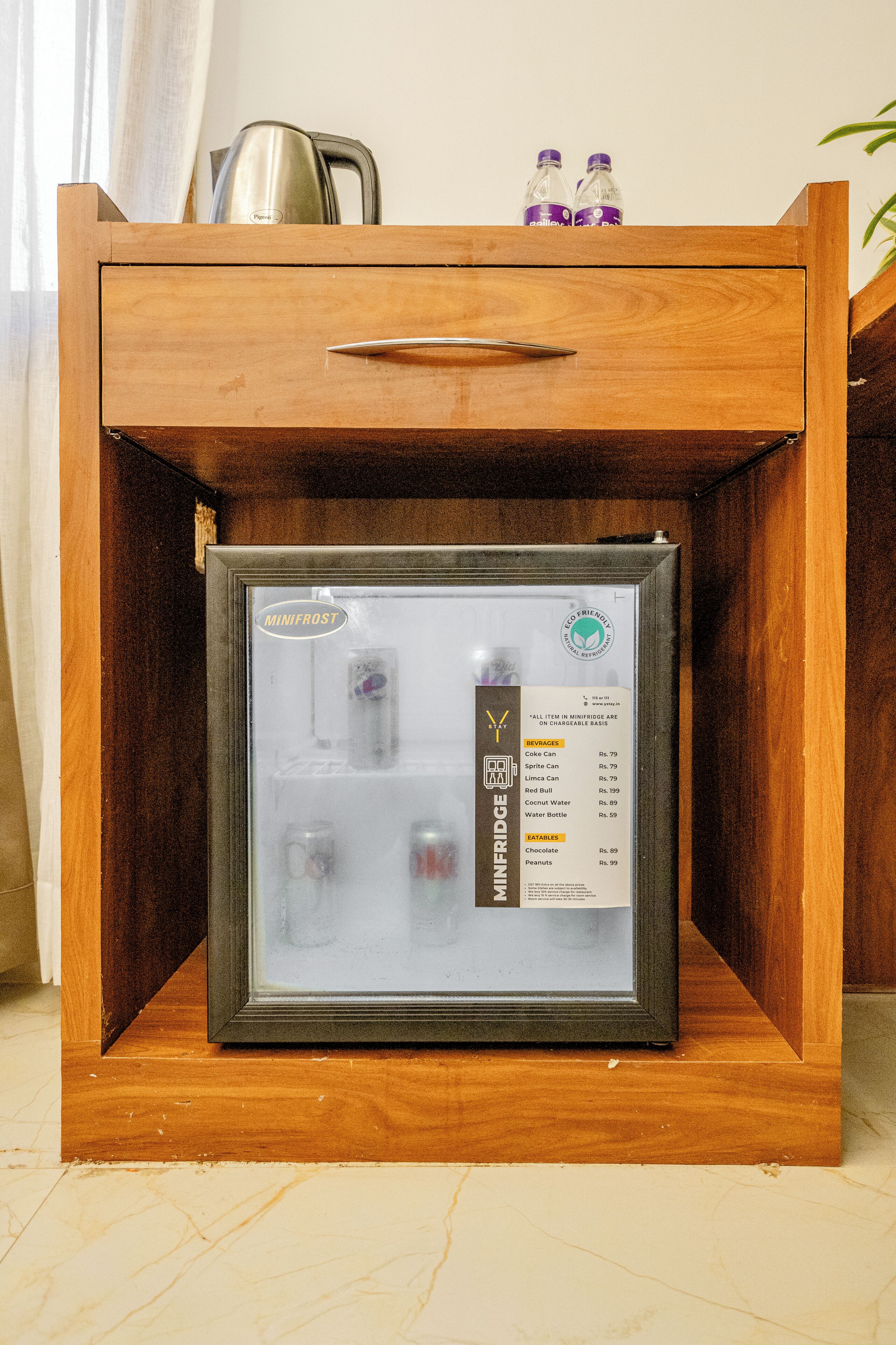Un minibár con puerta de vidrio, que contiene un refrigerador pequeño y una variedad de bebidas y aperitivos.