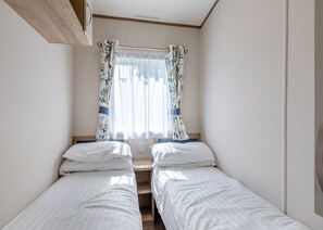 A small, clean room with two single beds, a window with floral curtains, and a wooden ceiling. - Gold 2 bedroom Caravan (Brixham, Torbay)