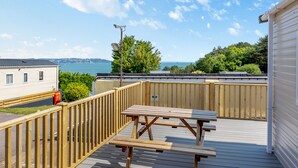 A deck with picnic tables overlooking a body of water, a white building, and a view of the sea. - Gold 2 bedroom Caravan (Brixham, Torbay)
