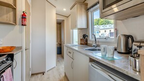 A compact kitchen with a microwave, stove, sink, and a window with a view of the outside. - Gold 2 bedroom Caravan (Brixham, Torbay)