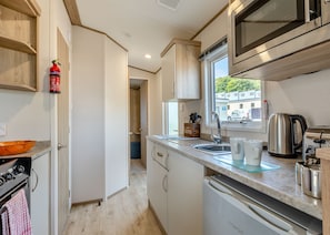 A compact kitchen with a microwave, stove, sink, and a window with a view of the outside. - Gold 2 bedroom Caravan (Brixham, Torbay)