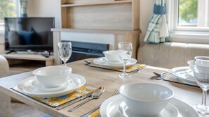 A dining table set with white plates, bowls, and wine glasses, with a television in the background. - Gold 2 bedroom Caravan (Brixham, Torbay)