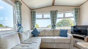 A cozy living area with a sectional sofa, a flat-screen TV, and large windows offering a view of the outdoors. - Gold 2 bedroom Caravan (Brixham, Torbay)