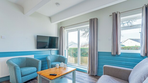 A room with a blue and white color scheme, a television mounted on the wall, a wooden table, and two sofas. - Shoreline Apartment 3 (Pets Welcome) (Woolacombe)