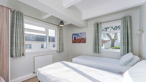 A bedroom with two beds, a radiator, a window with curtains, a painting, and a ceiling light. - Shoreline Apartment 3 (Pets Welcome) (Woolacombe)