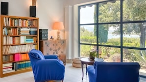 Une pièce avec une grande fenêtre, une bibliothèque, deux chaises bleues, une petite table avec une lampe et un radiateur.