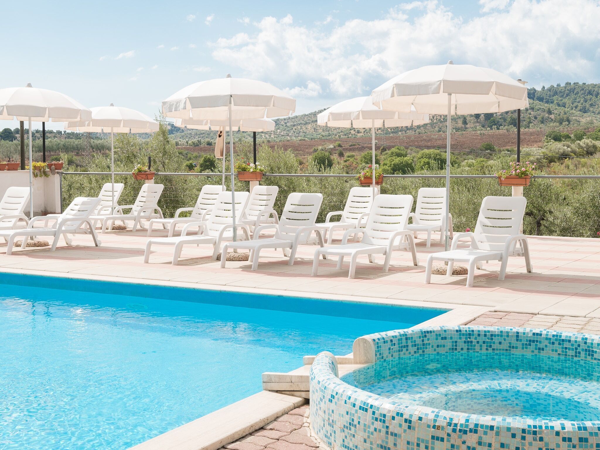 A pool area with white lounge chairs and umbrellas, a tiled hot tub, and a view of a hilly landscape.