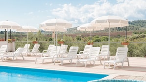 Un espace piscine doté de chaises longues blanches et de parasols, d’un jacuzzi carrelé et offrant une vue sur un paysage vallonné.