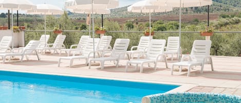 A pool area with white lounge chairs and umbrellas, a tiled hot tub, and a view of a hilly landscape.