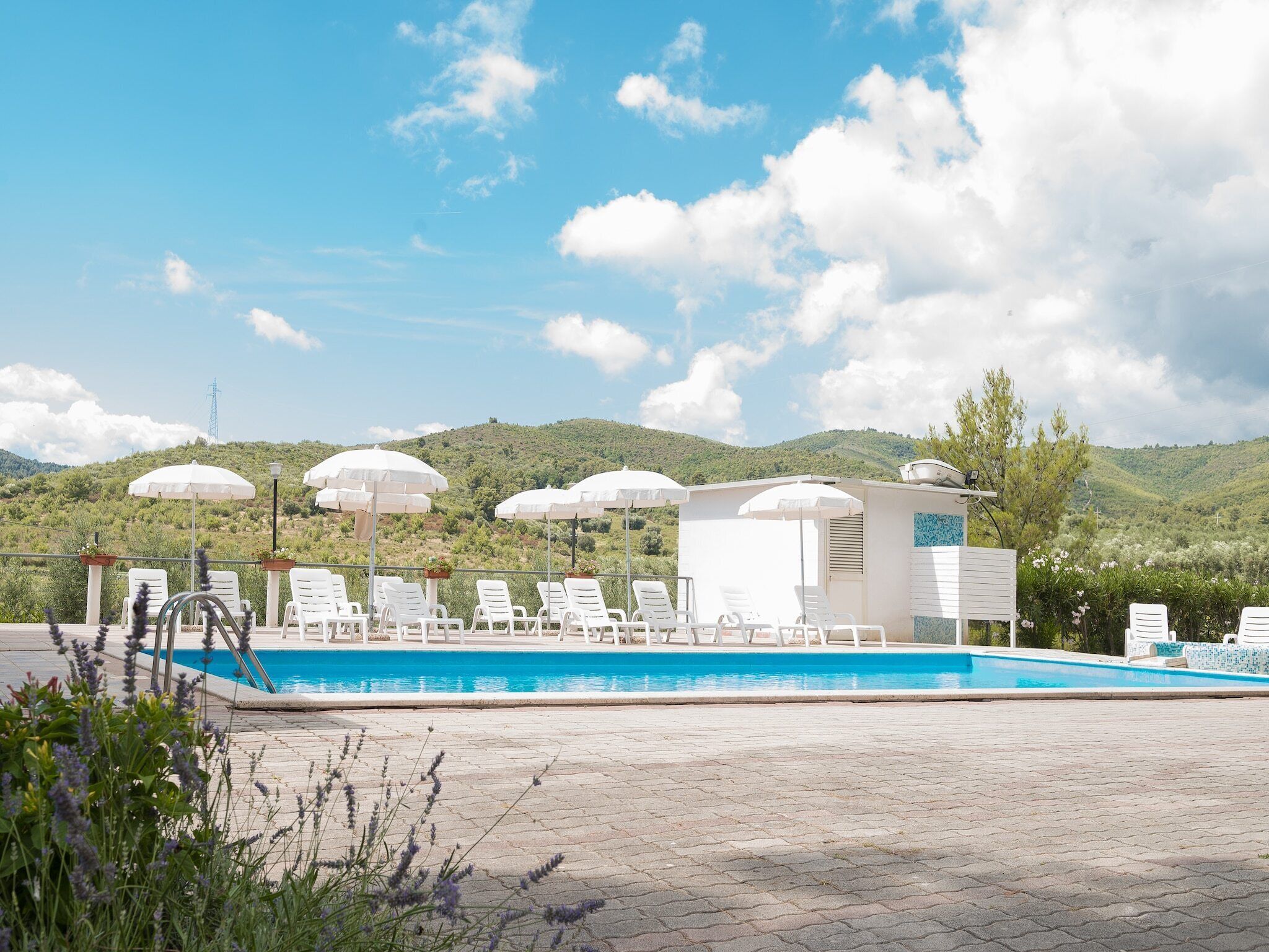 Zona de piscina con sillas reclinables blancas y sombrillas, rodeada de vegetación y colinas.