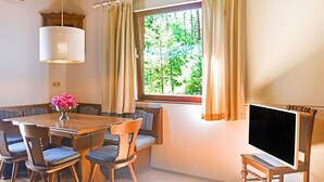 Ein Essbereich mit einem Holztisch, gepolsterten Stühlen, einer Chandelüster, einem Fernseher auf einem Holzständer und einem Fenster mit Vorhängen.