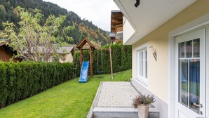 A residential area with a wooden swing set, a blue slide, and a paved pathway leading to a house with a white exterior and a window with a curtain.