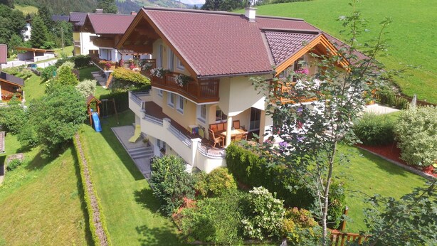 A multi-story house with a red-tiled roof, a balcony with wooden railings, and a well-maintained garden with various plants and a small play area.