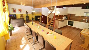 Une cuisine spacieuse avec un plafond en bois, un coin repas doté d’une longue table et un salon agrémenté d’une cheminée.