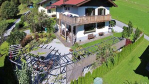 Un chalet a due piani con un balcone, immerso nel verde e circondato da montagne.