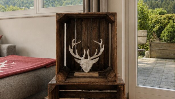 Un meuble rustique en bois, orné d’une tête de cerf, placé près d’une fenêtre donnant sur une colline boisée.