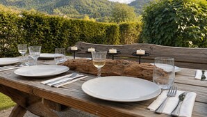 Une table à manger en bois, installée à l’extérieur, avec des assiettes, des verres et des bougies, offrant une vue sur un paysage montagneux.