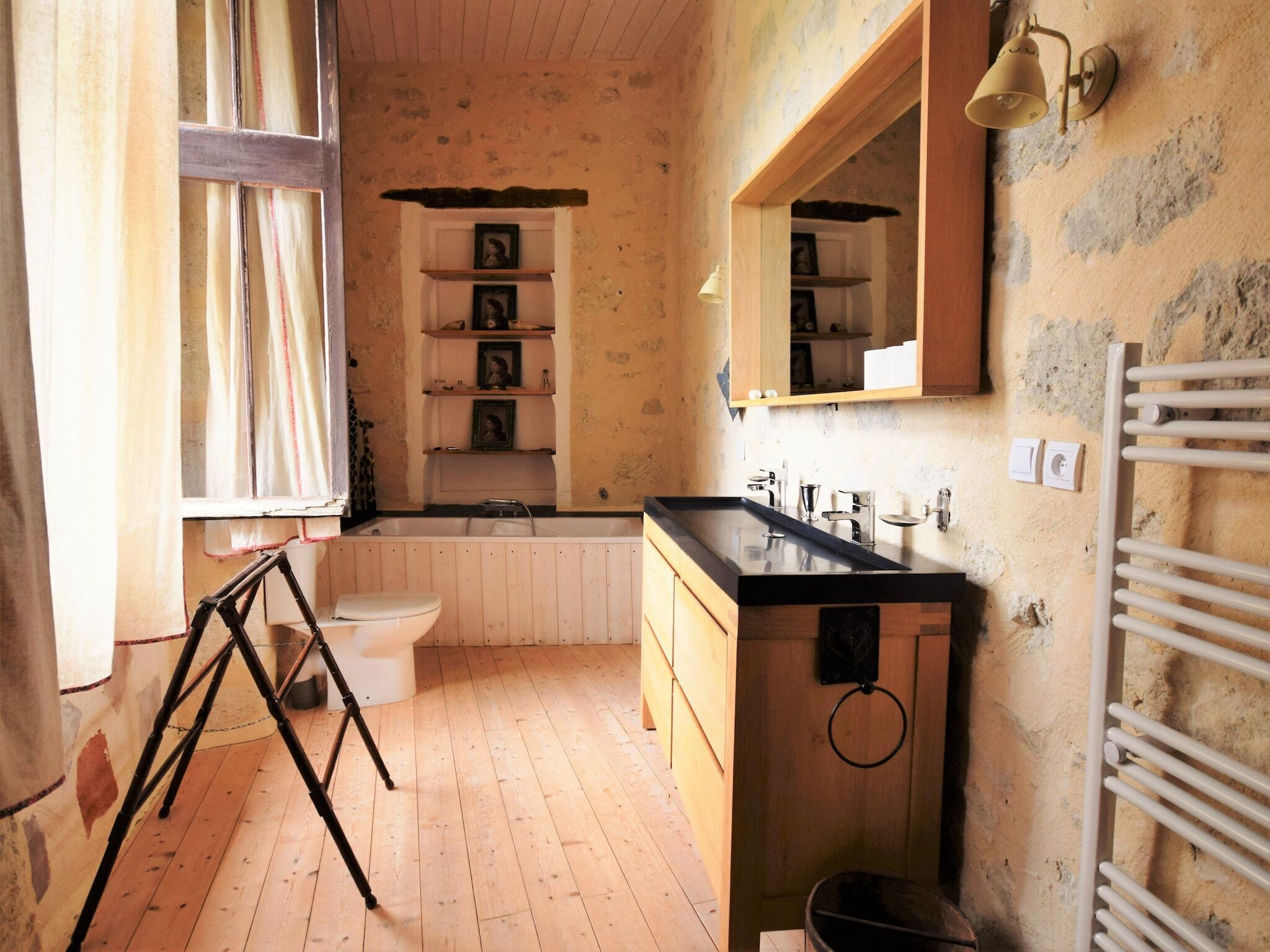 A rustic bathroom with wooden floors, a white toilet, a black sink, and a mirror above the sink. Shelves with framed pictures are mounted on the wall.